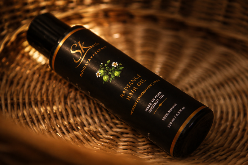 A bottle of SK Sundara Kayakalp Skincare Radiance Hair Oil resting on a woven wicker tray under warm, golden lighting.